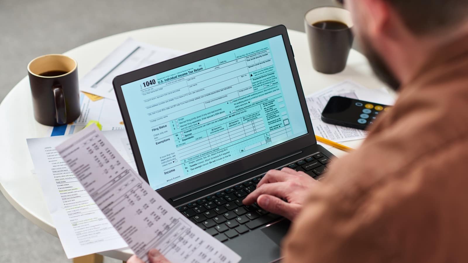 A middle-aged man filing forms online at a laptop, surrounded by financial documents