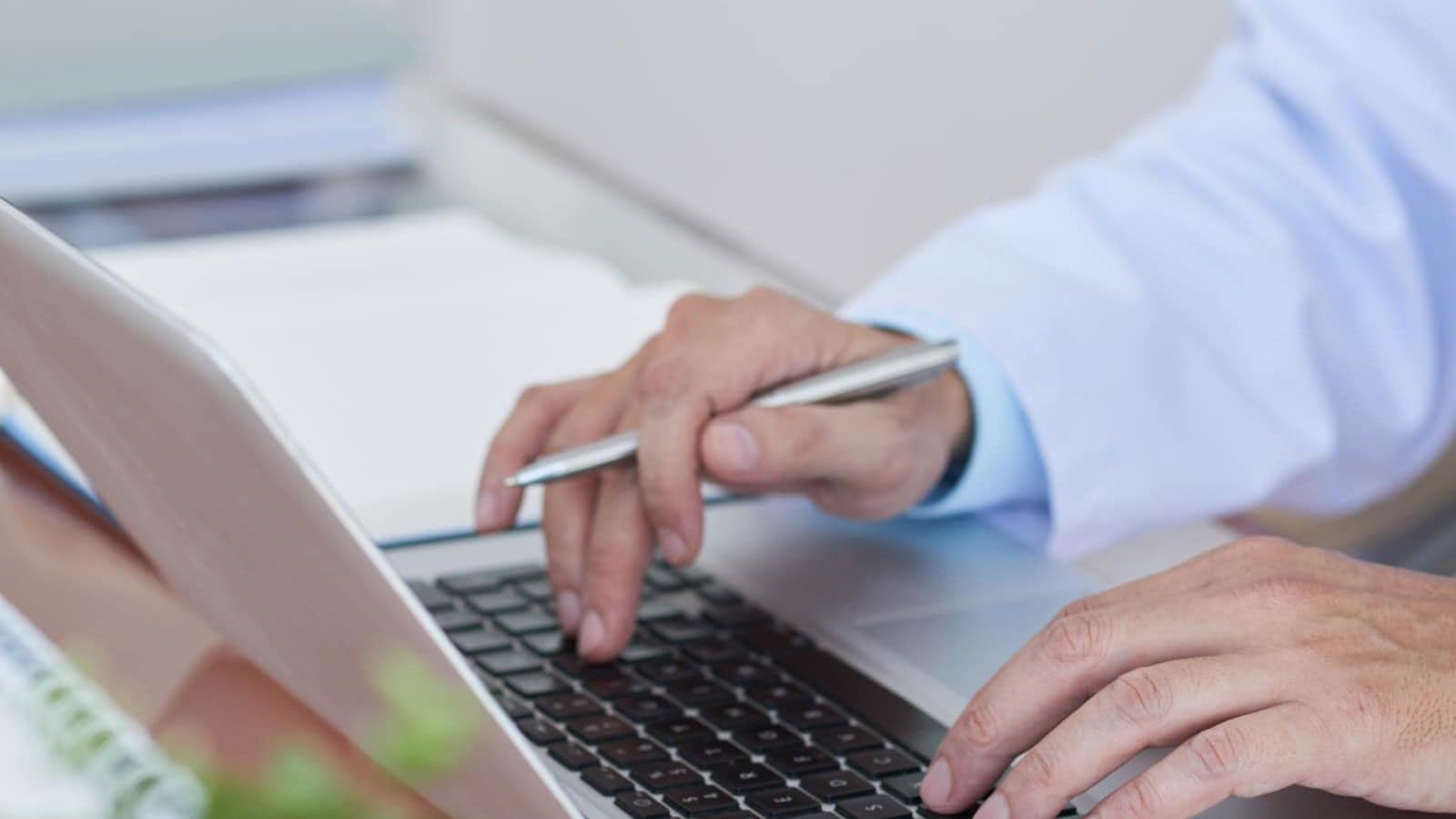 Close-up of a clinician's hands typing on a laptop while holding a pen, with paper records open alongside