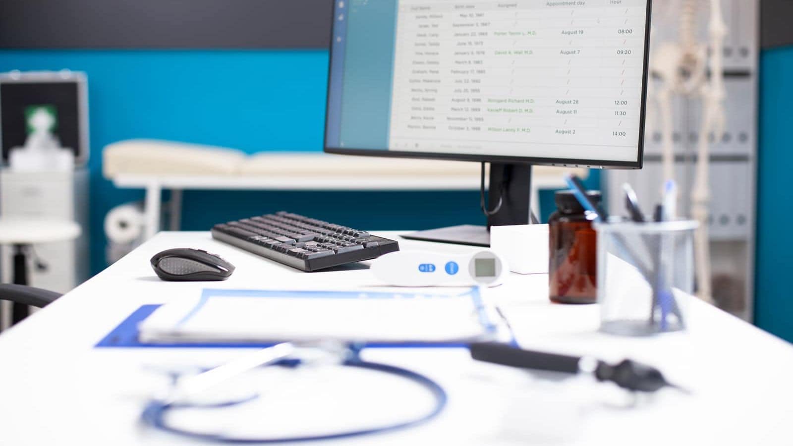An unattended clinic desk, a monitor showing a patient scheduling list beside a stethoscope, thermometer and clipboard