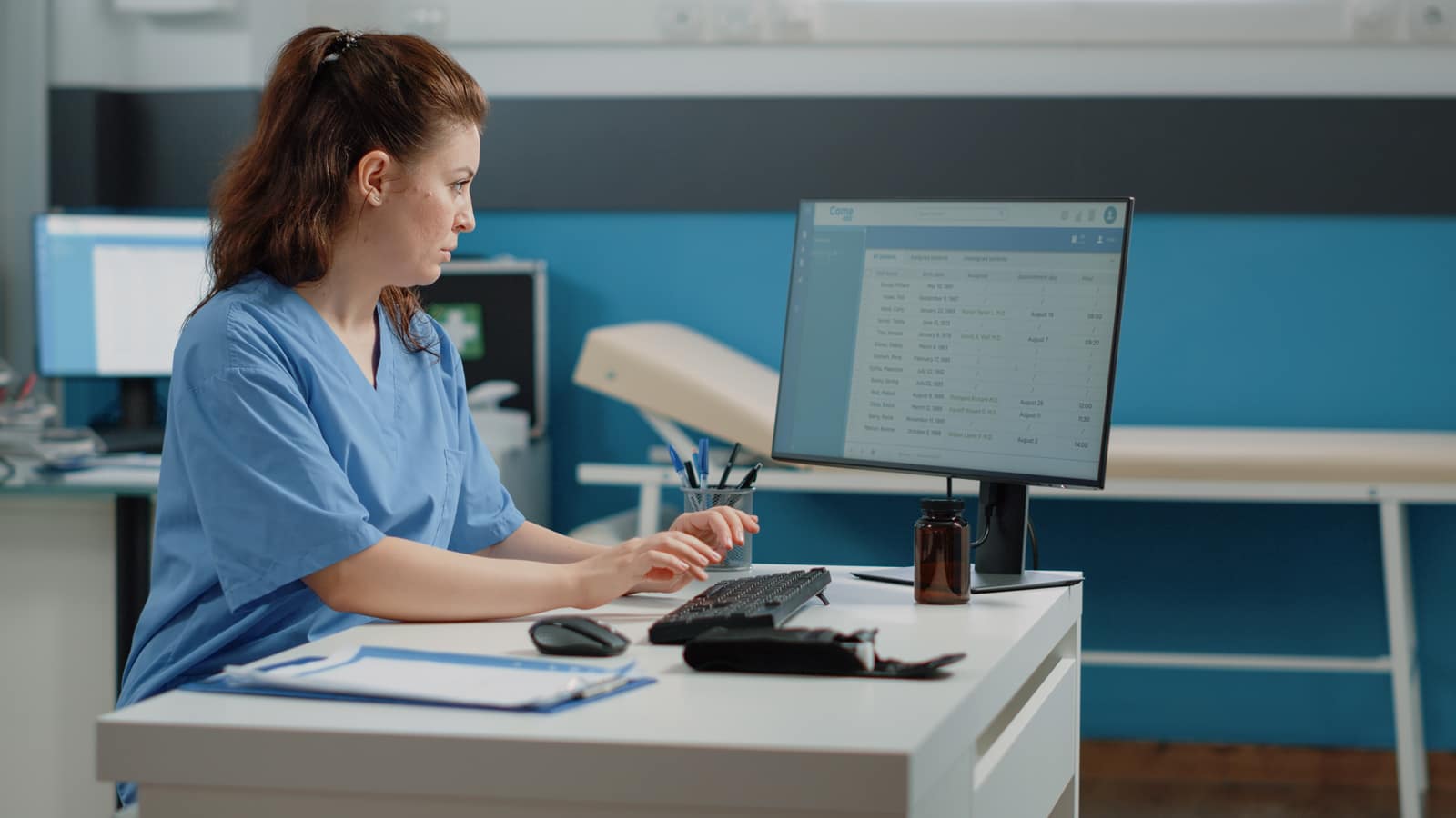 A nurse in blue scrubs typing at a clinic workstation, a patient list with dates and appointment times on the monitor